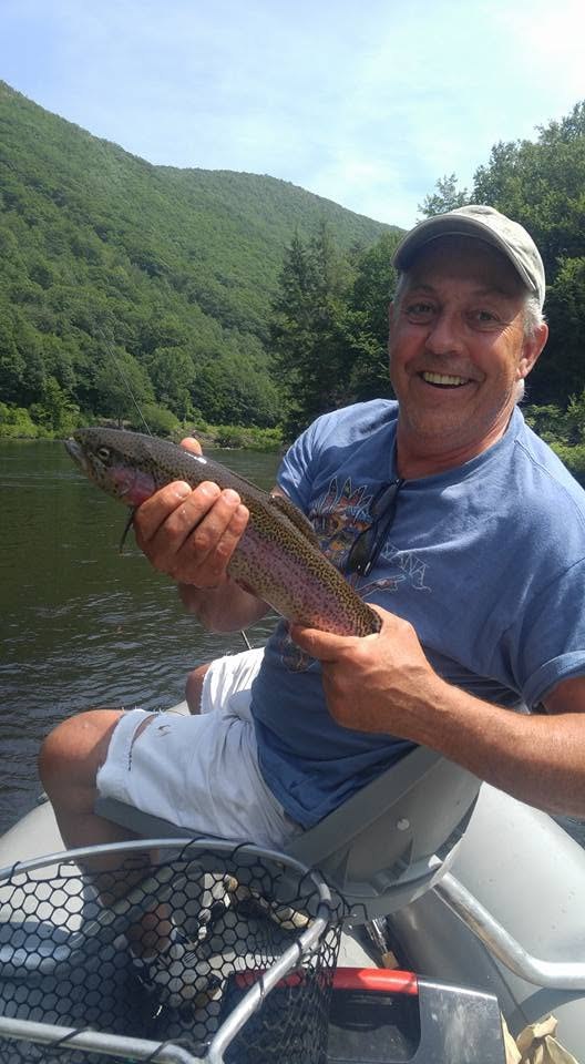 Paul King sitting in a boat on a river, holding a trout, with forested hills behind him.