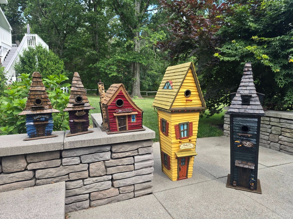 Five hand-painted birdhouses lined up on a stone garden wall: a blue house, a purple house, a red cottage, a tall yellow house, and a dark green tavern.